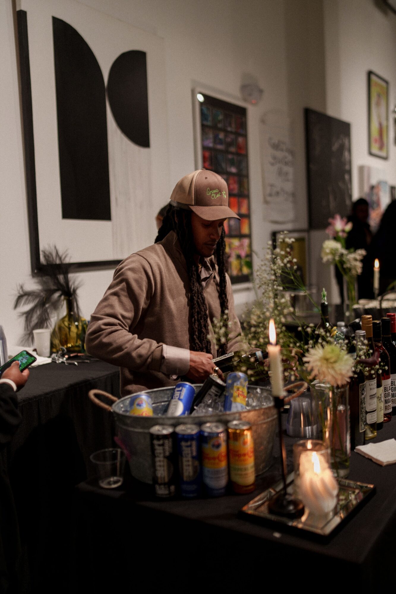 Person wearing a cap stands behind a table of drinks, flowers, and candles in a gallery-like room.