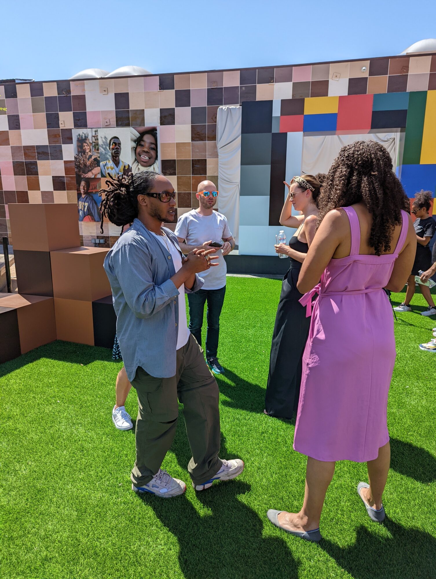 People talk on a lawn in front of a colorful tiled wall and geometric display.