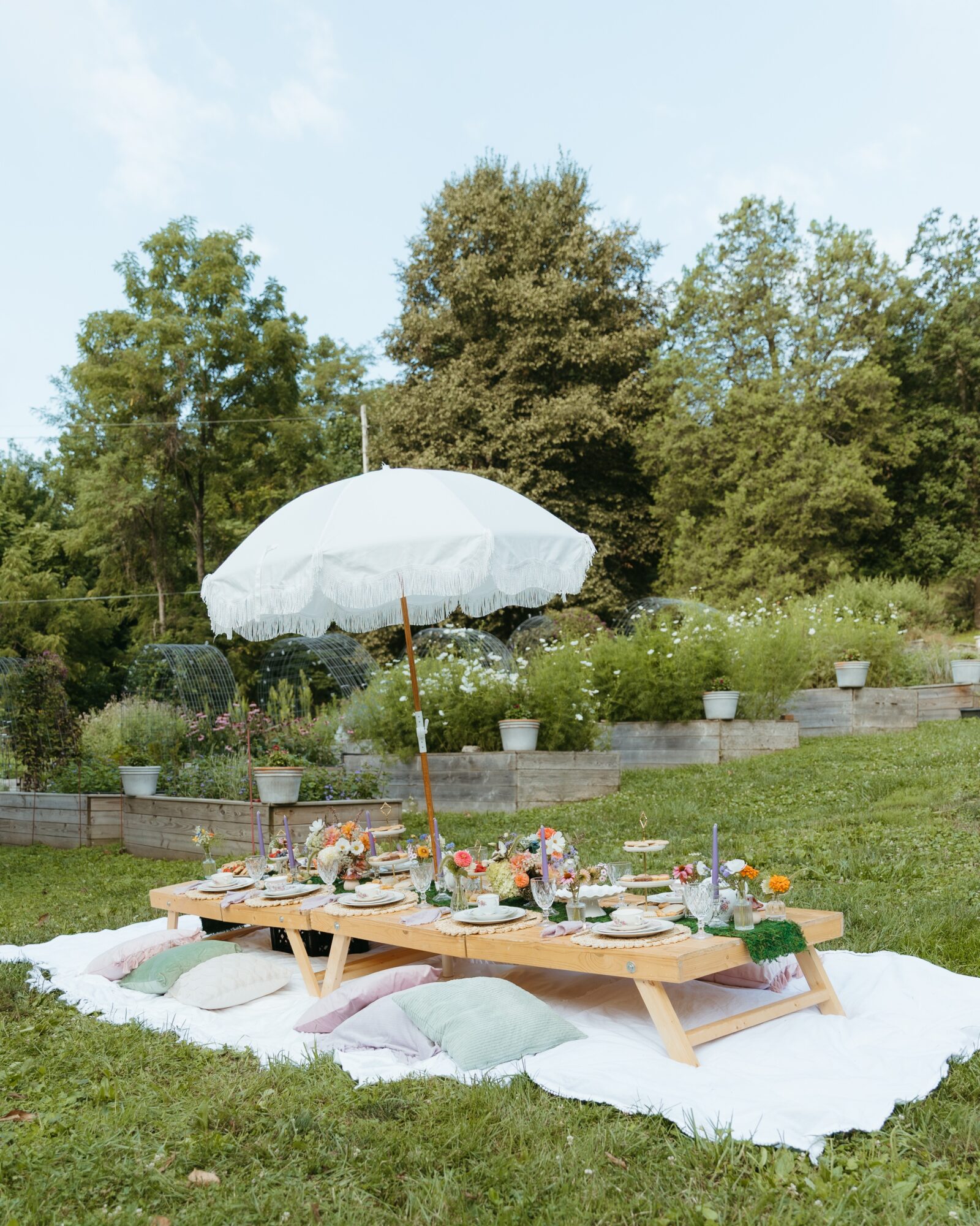 A picnic table with food and flowers sits on a blanket beneath a white umbrella in a garden.