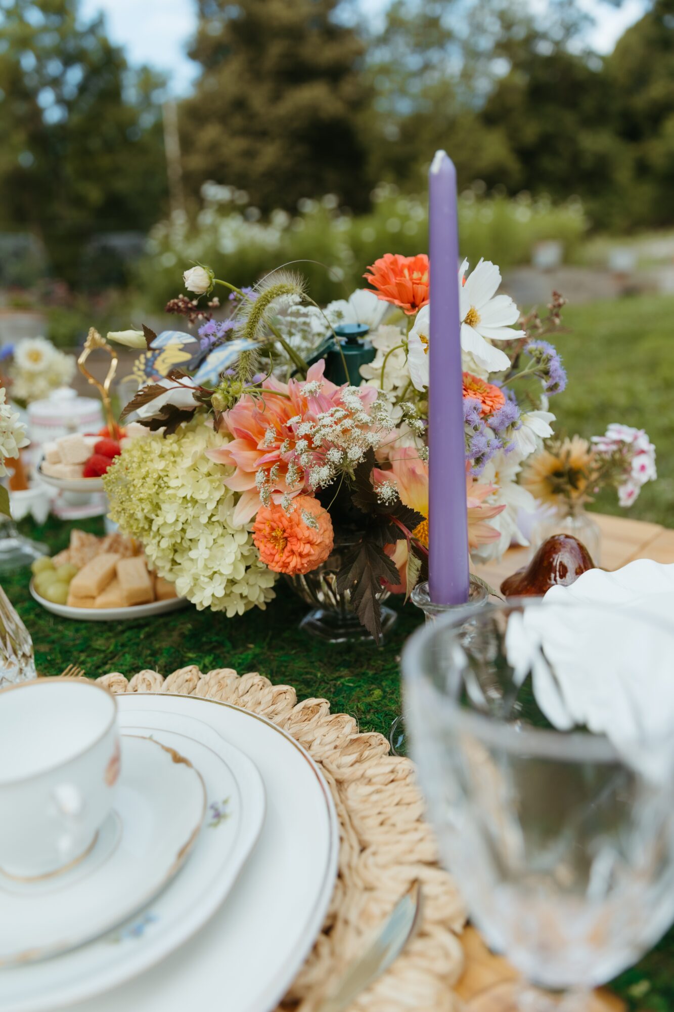 A purple taper candle stands among flowers on a table set with white dishes and glassware outdoors.