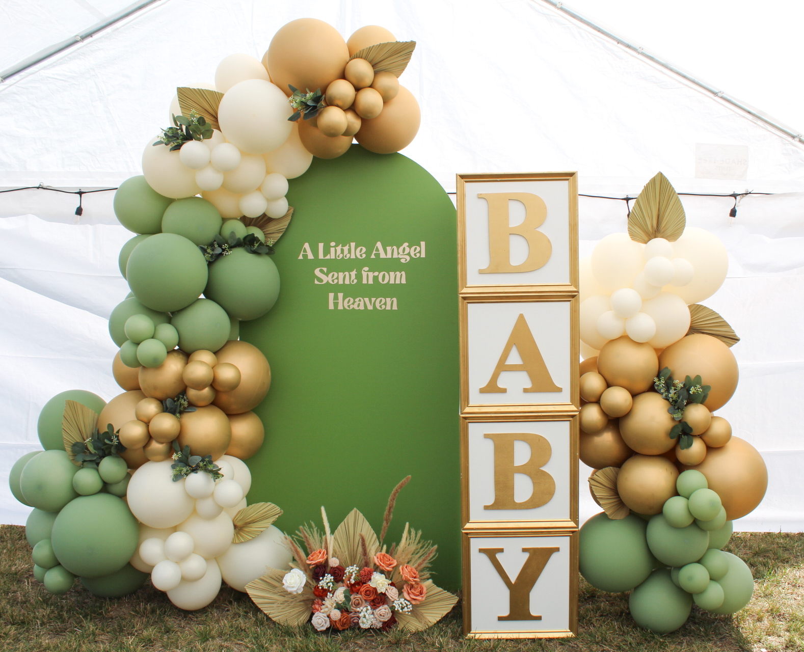 Green, white, and gold balloon arch beside stacked blocks spelling BABY, with a sign reading “A Little Angel Sent from Heaven.”