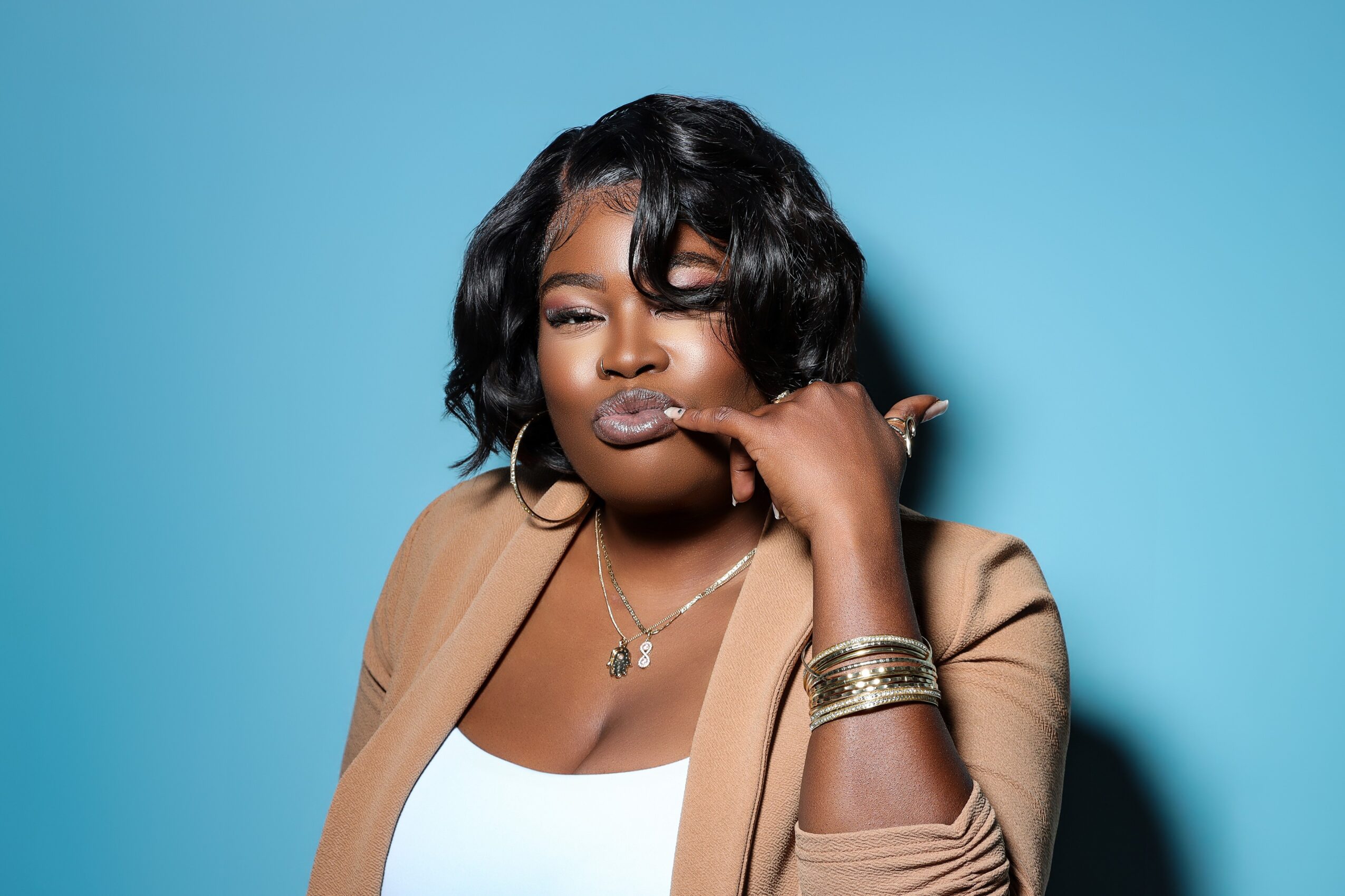 Person with dark hair wearing a tan blazer and gold jewelry against a blue background.