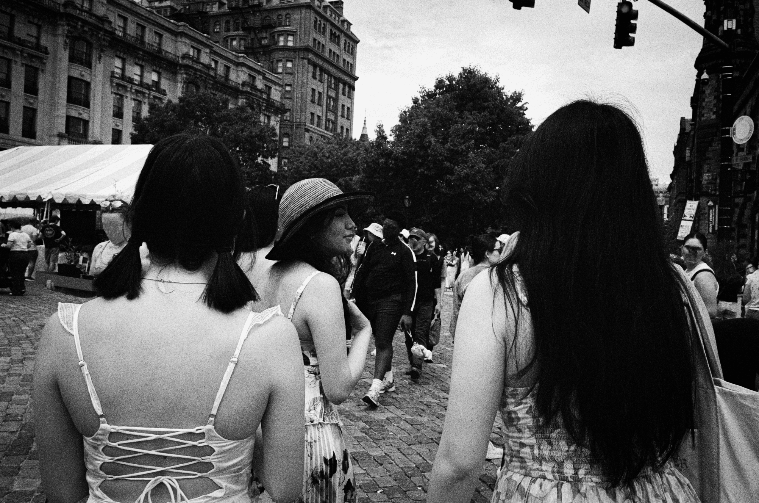 Three people walk through a crowded city street, with buildings and a market tent nearby.