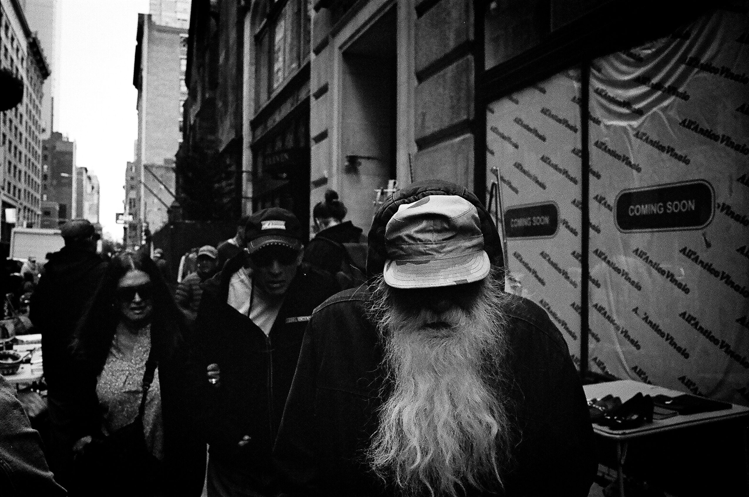 Black-and-white city street scene with pedestrians, including an older person with a long white beard and brimmed hat.