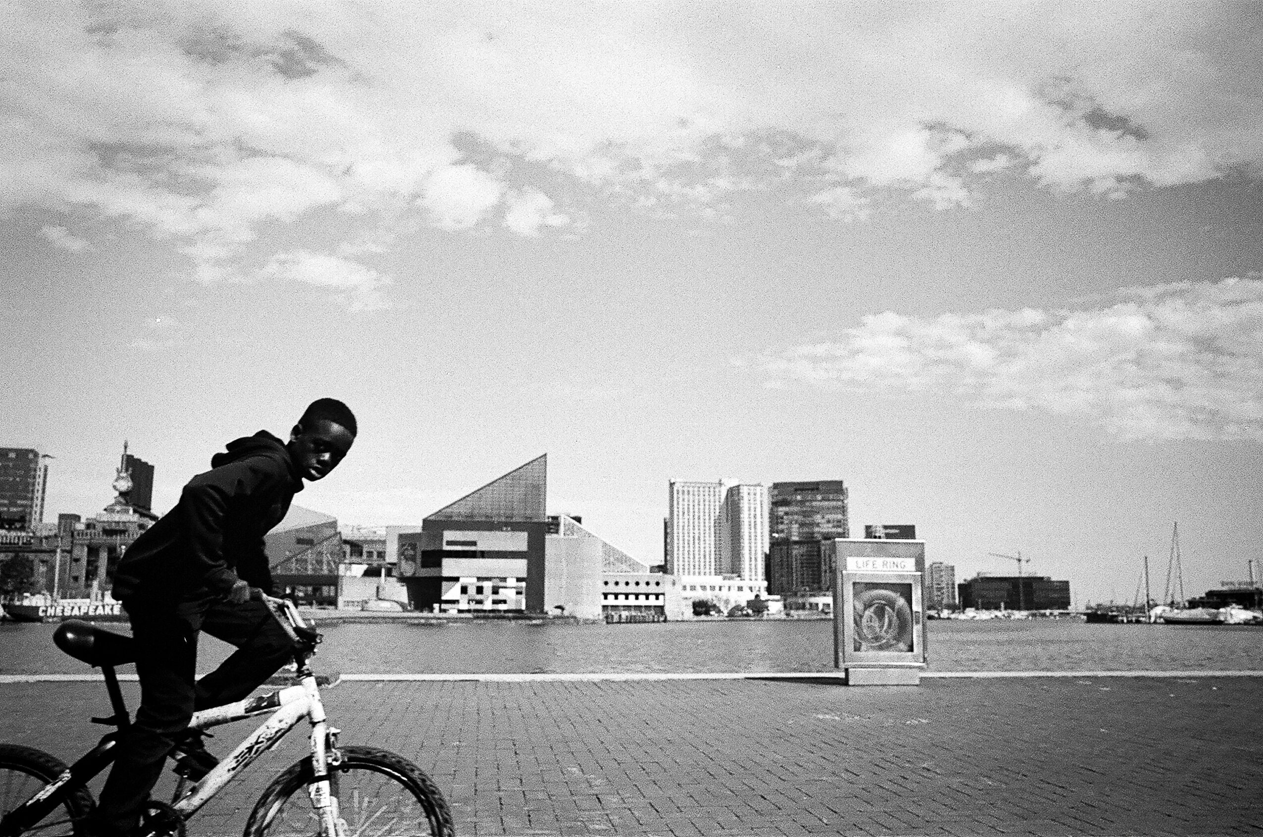 A person crouches on a bicycle along a waterfront promenade, with city buildings and a cloudy sky behind them.
