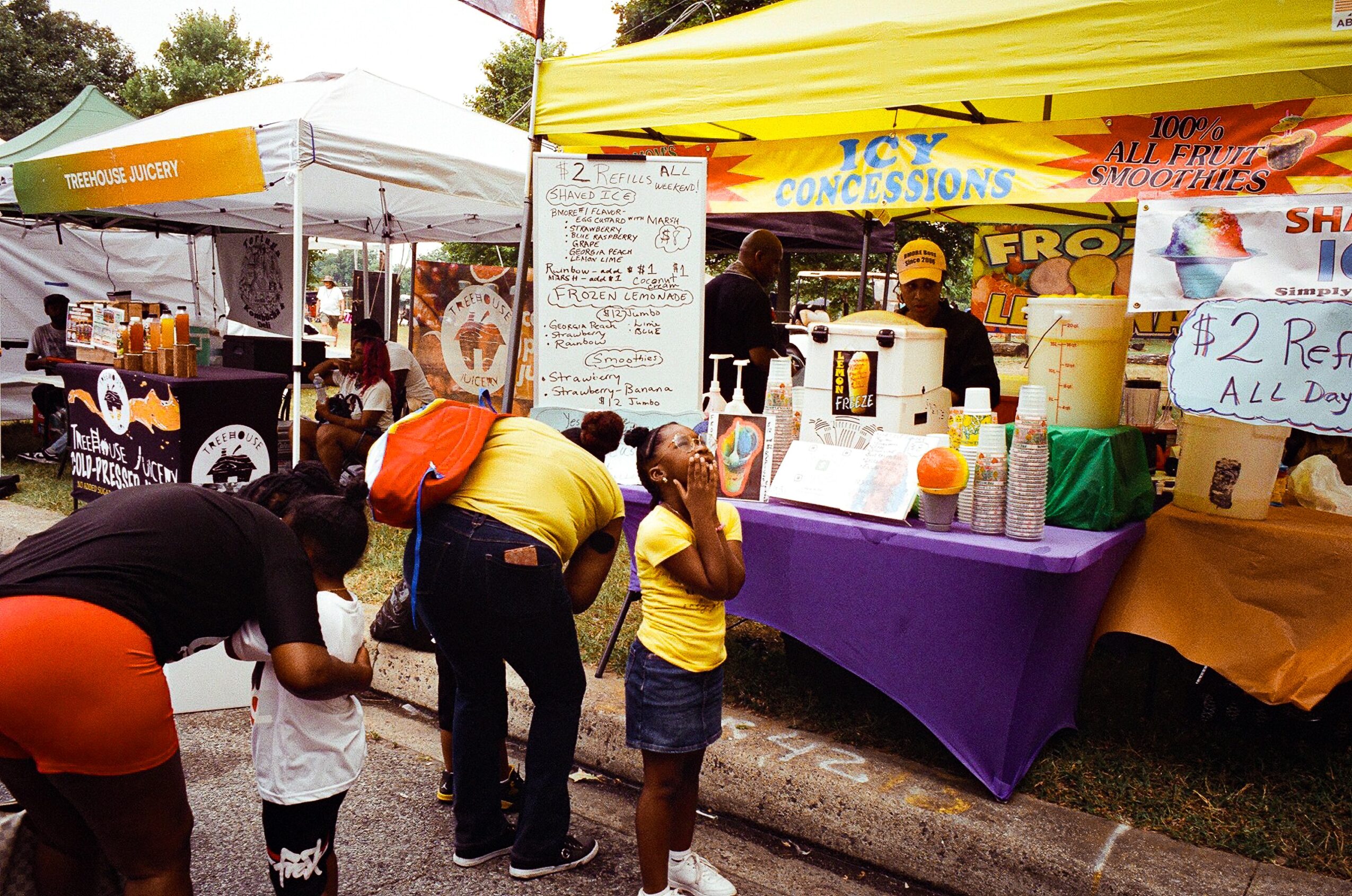 People browse a concession stand at an outdoor market, with colorful signs, food containers, and a purple-covered table.