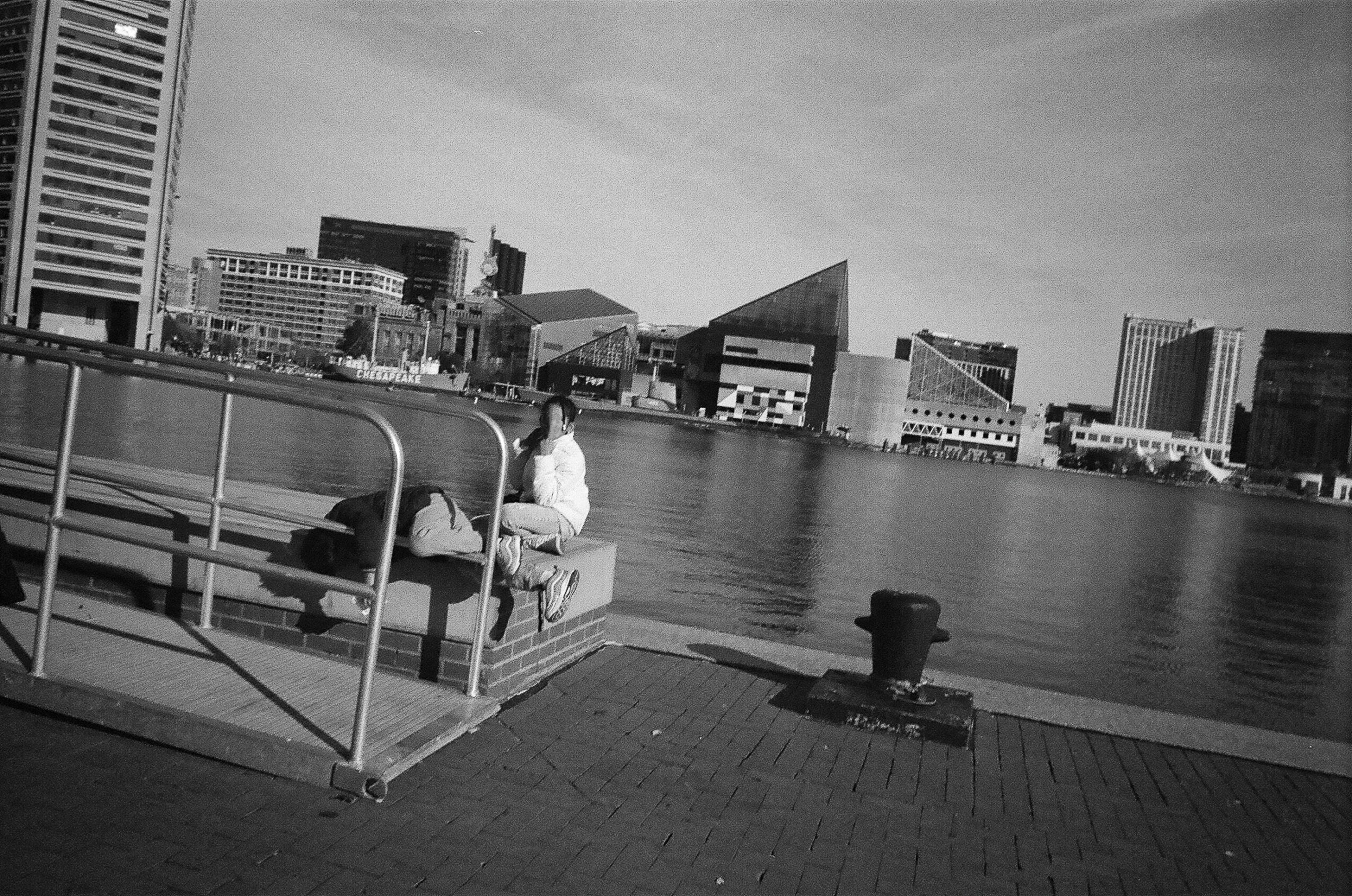 Black-and-white waterfront scene with a seated person, railing, paved promenade, and city buildings across the water.