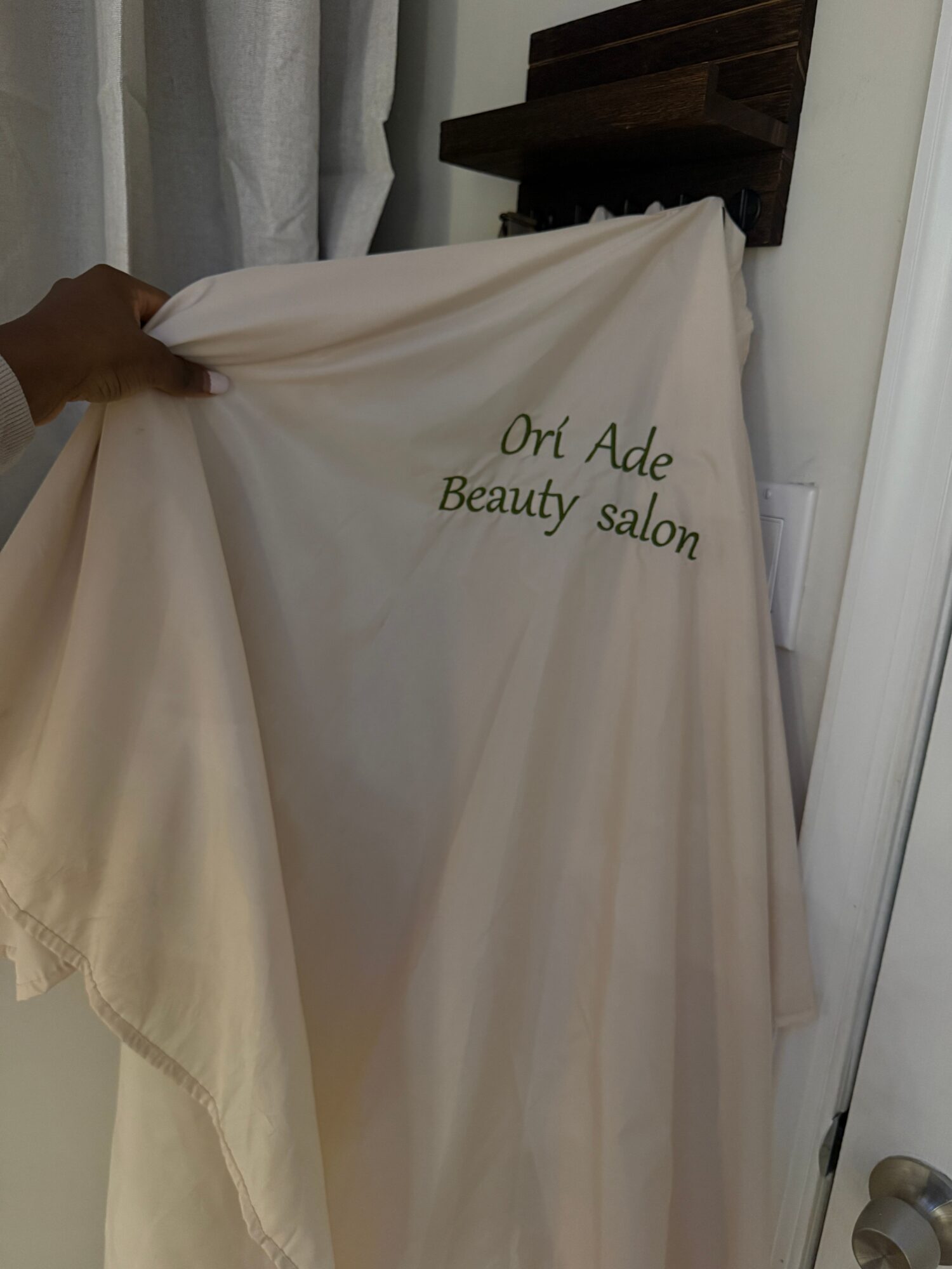 A hand holds the edge of a light-colored fabric curtain embroidered with “Our Ade Beauty Salon.”