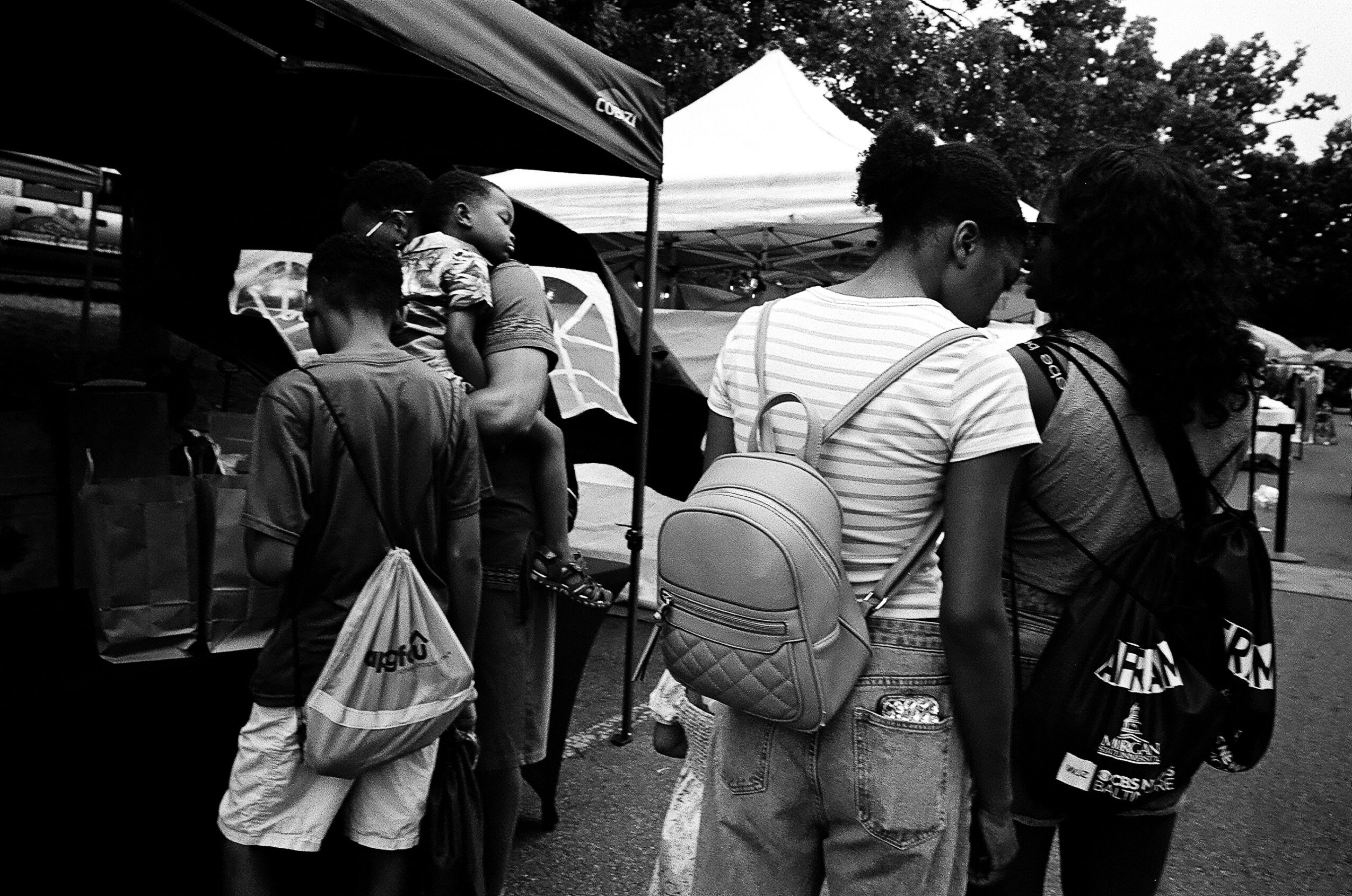 People browse a market stall beneath canopy tents, with one person carrying a child and another wearing a backpack.