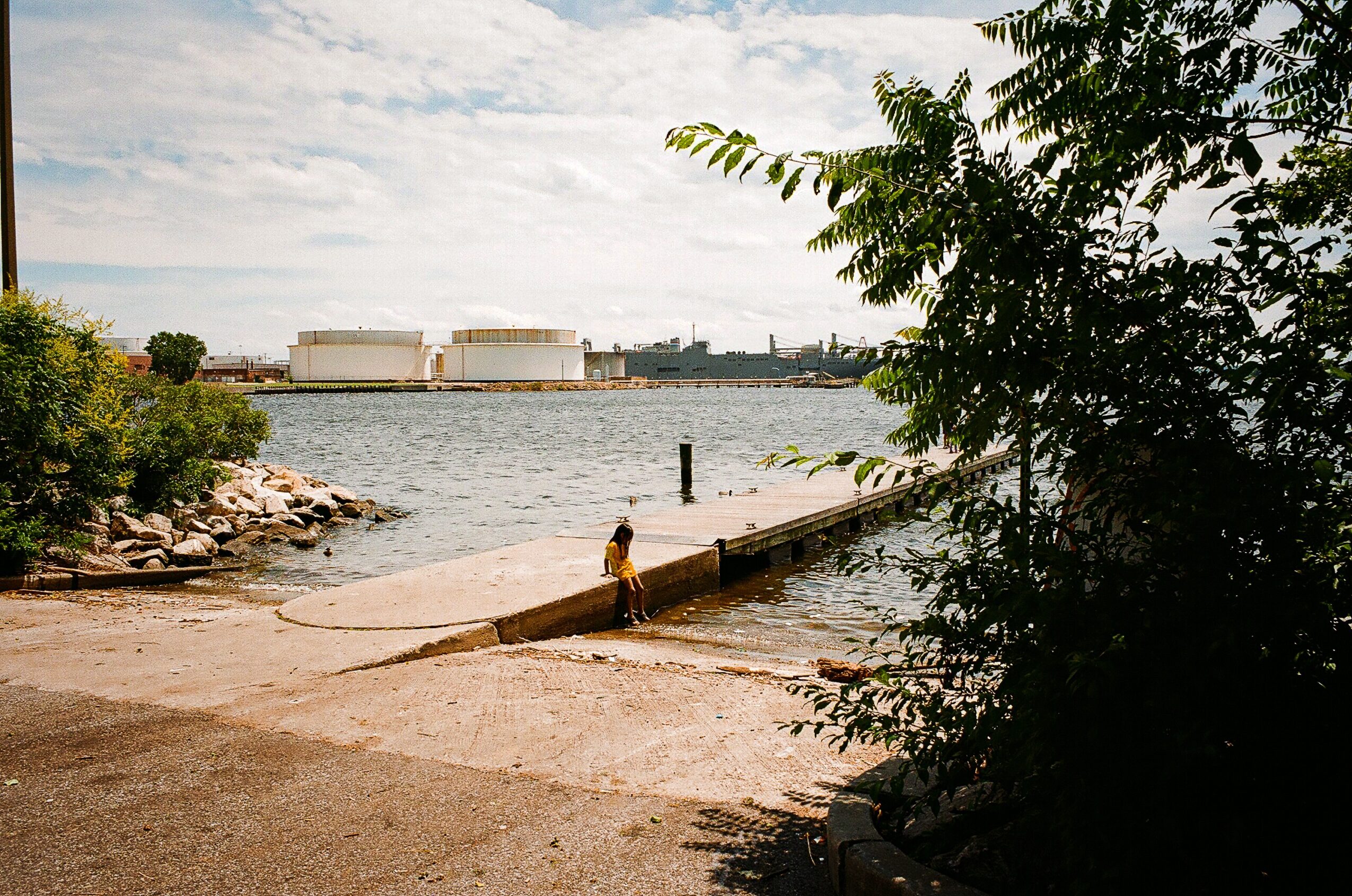 A person stands on a sloped concrete ramp beside calm water, with trees framing the view and industrial tanks across the shore.