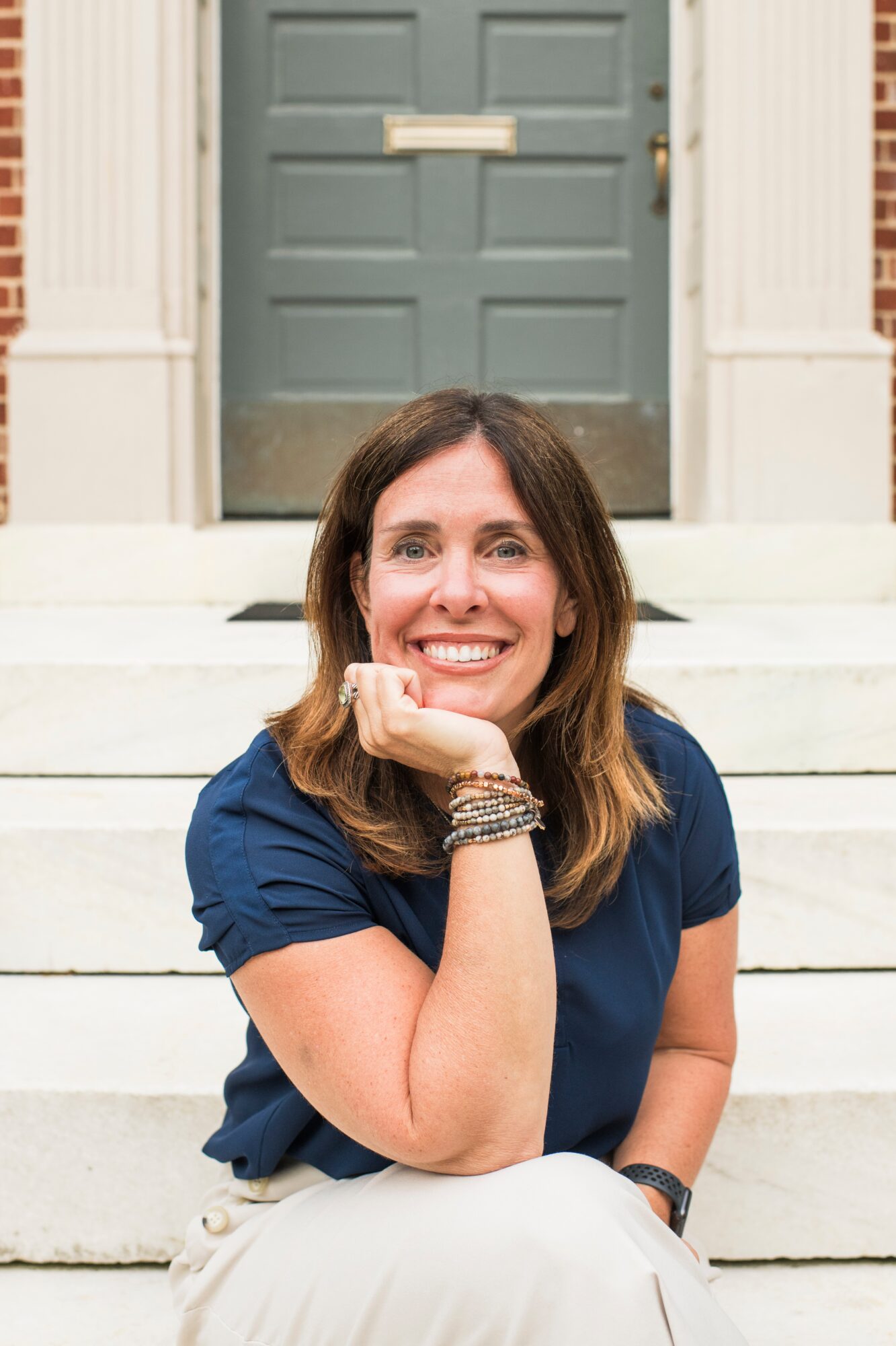 A person with brown hair sits on stone steps in front of a gray door, resting her chin on one hand.