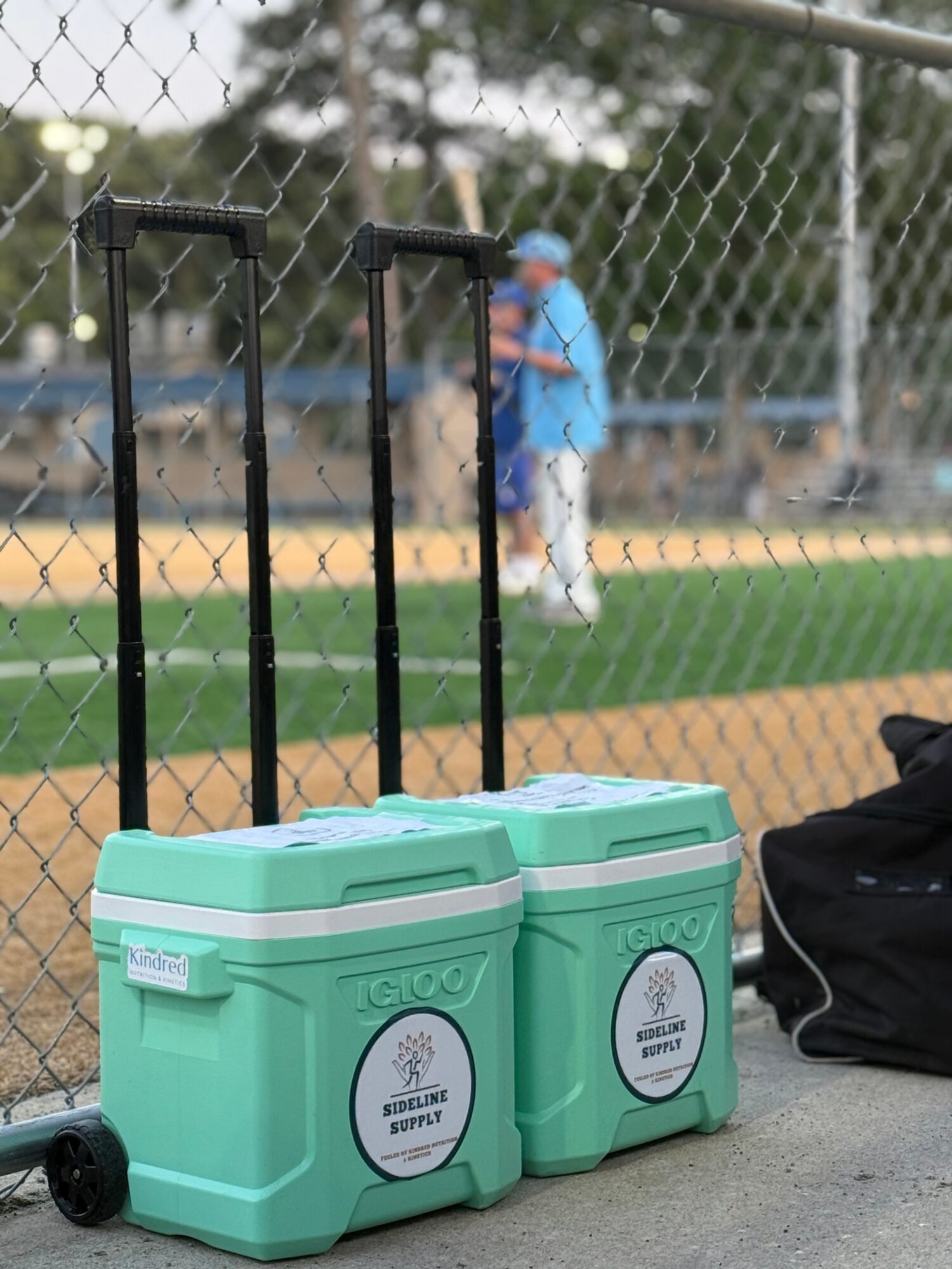 Two green coolers sit beside a chain-link fence, with a person on a baseball field in the background.