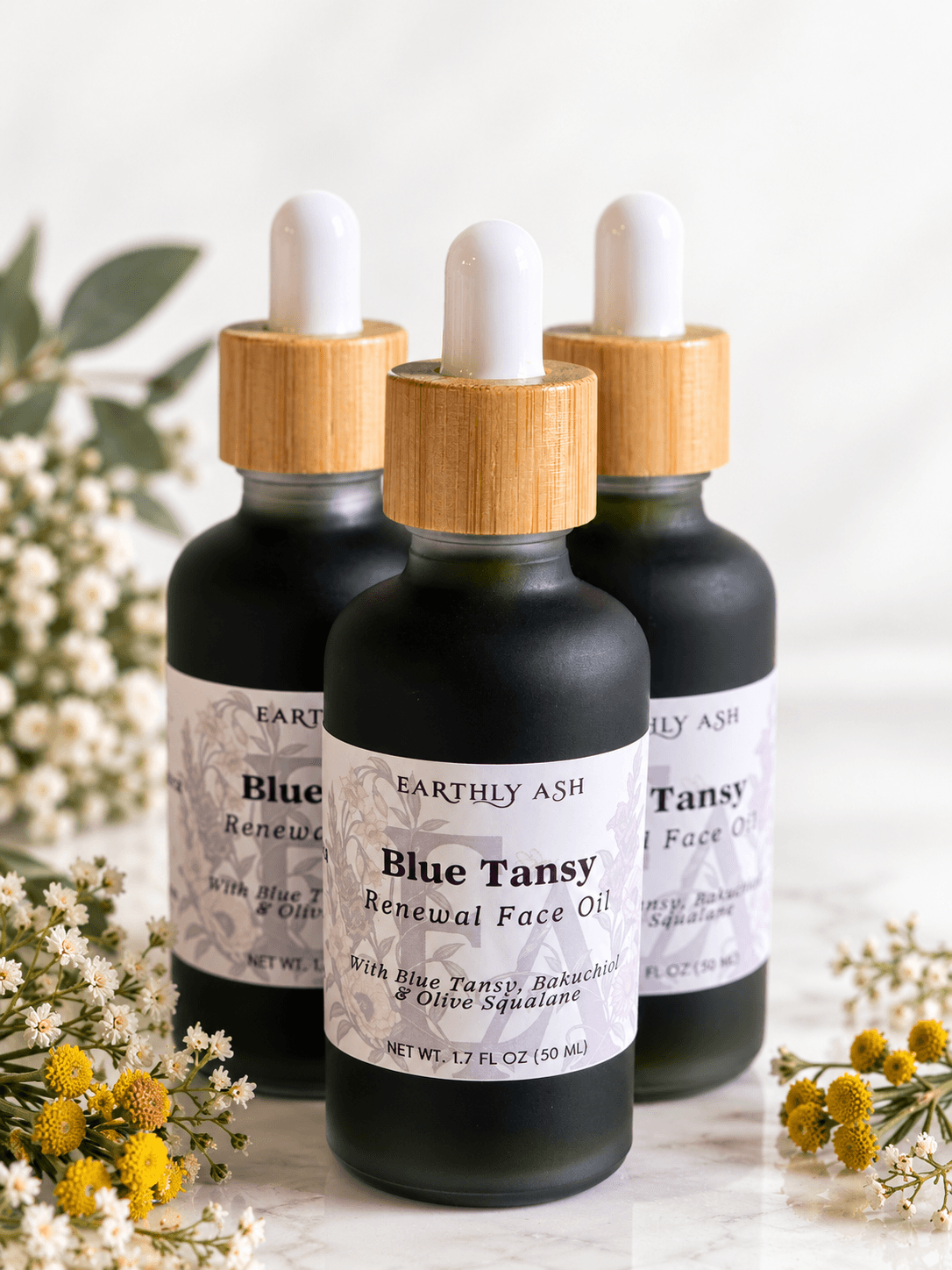 Three dark glass dropper bottles with wooden caps and labels among small white and yellow flowers.