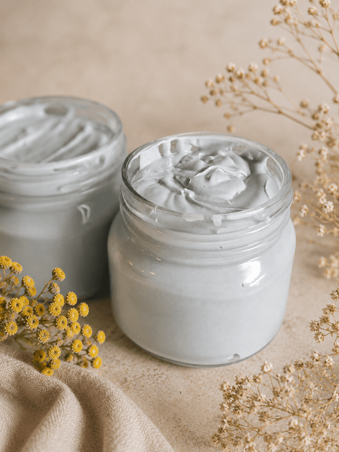 Two open glass jars of white cream sit among dried yellow flowers and beige fabric.