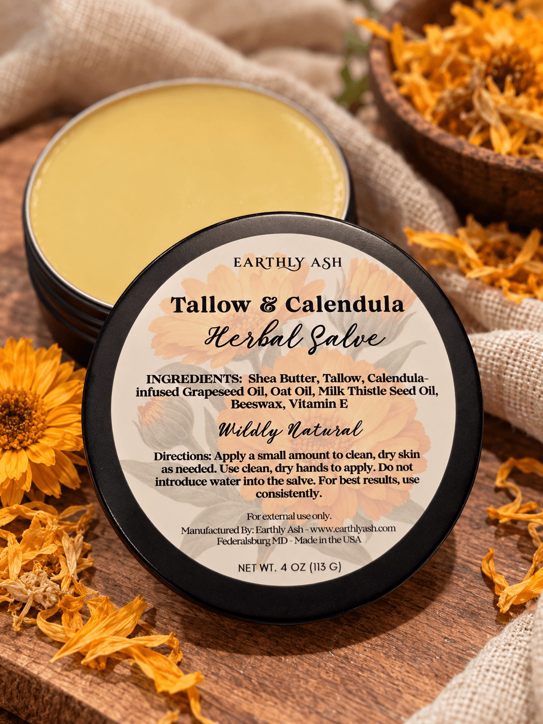 Two round tins of yellow herbal balm sit on a wooden surface beside dried orange flowers and beige fabric.