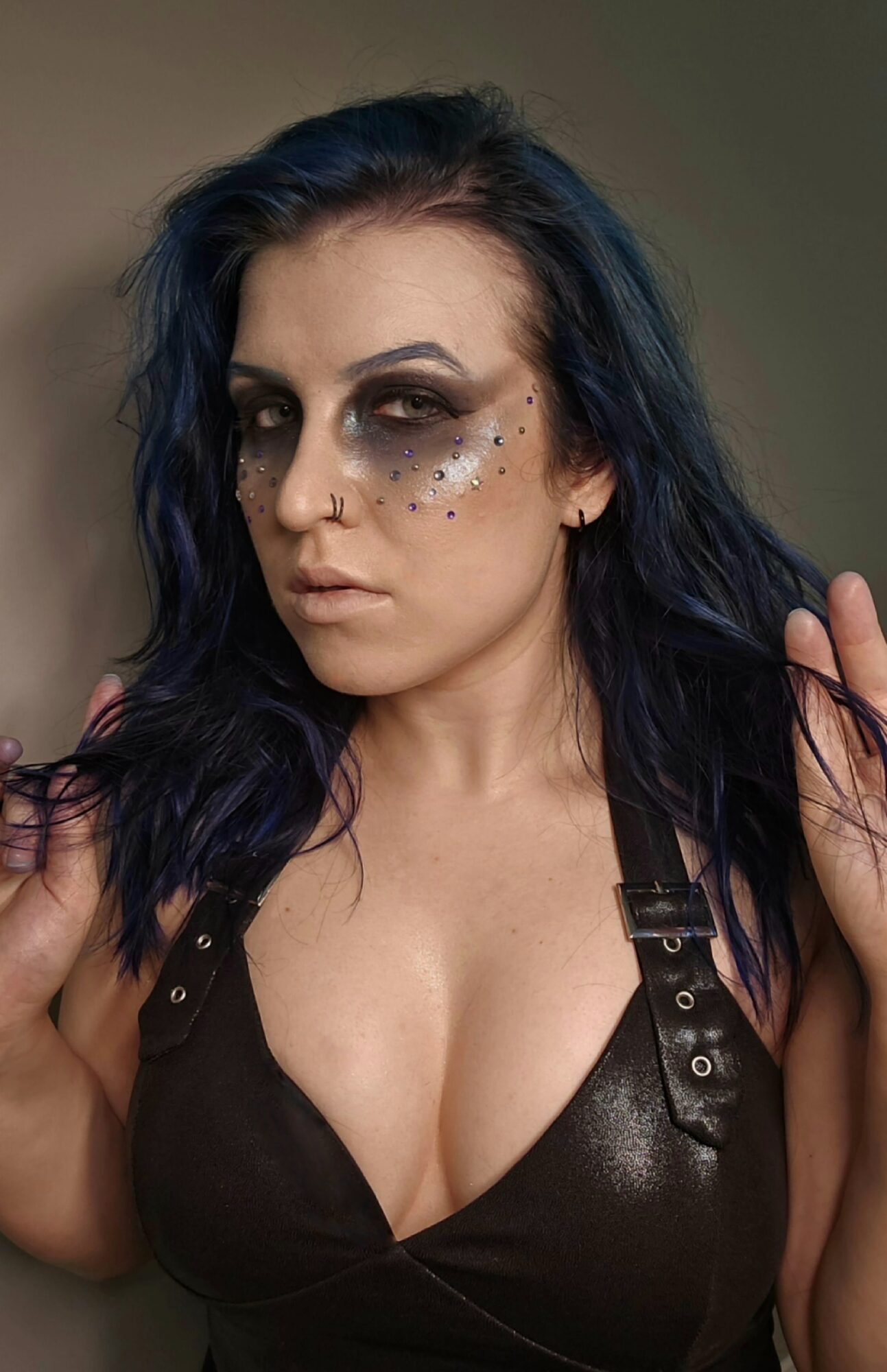 Person with long dark hair, dramatic eye makeup, and a black leather top against a plain background.