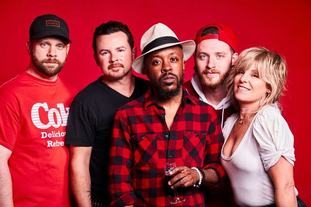Five people pose against a red background; the central person wears a red-and-black checkered shirt and white hat.