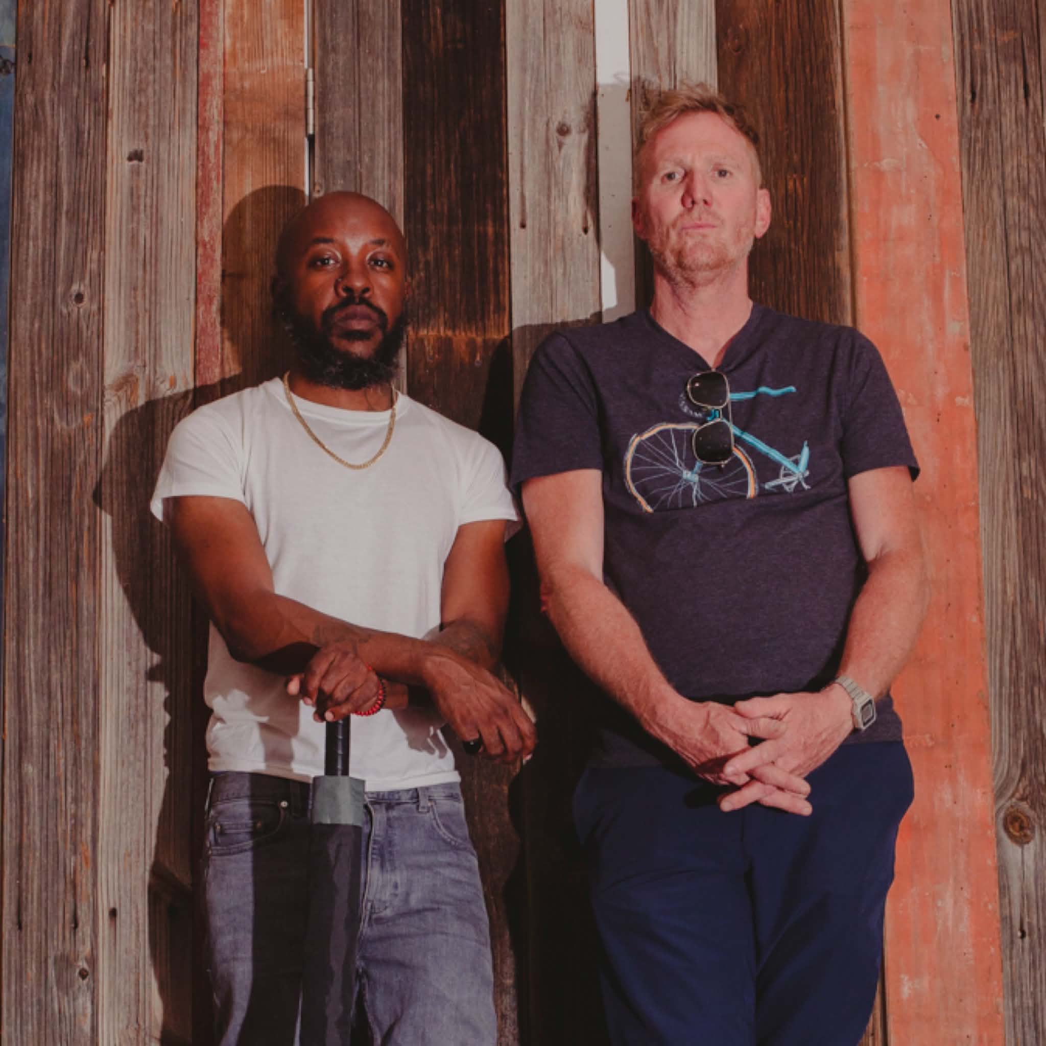 Two people stand against a weathered wooden wall; one holds a closed umbrella.