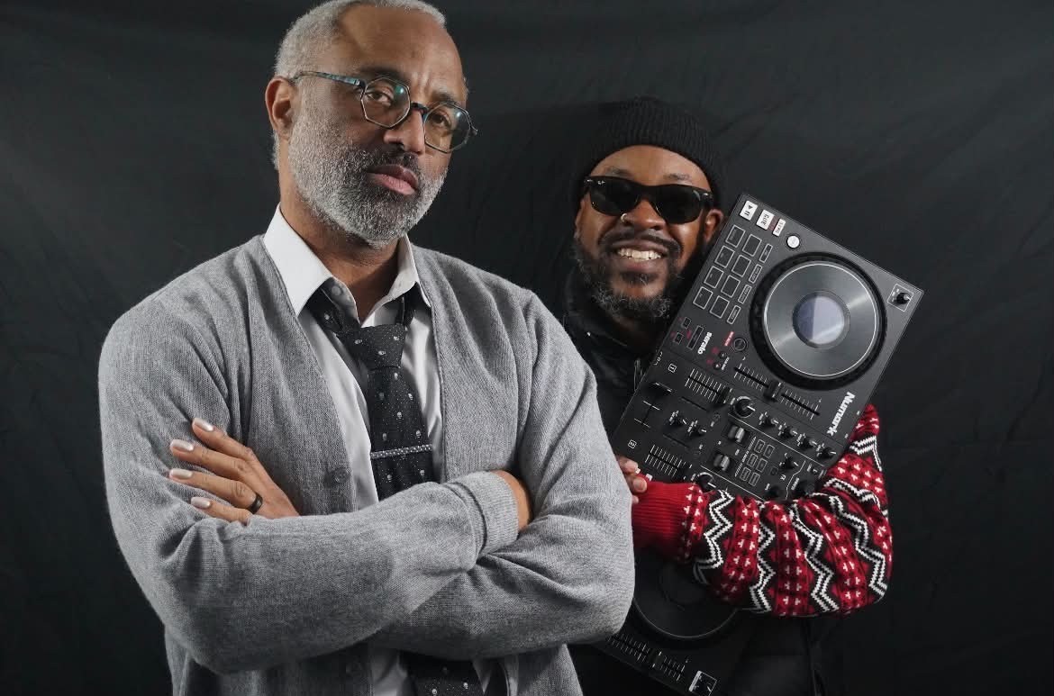 Two people pose against a dark background; one crosses his arms while the other holds DJ equipment.