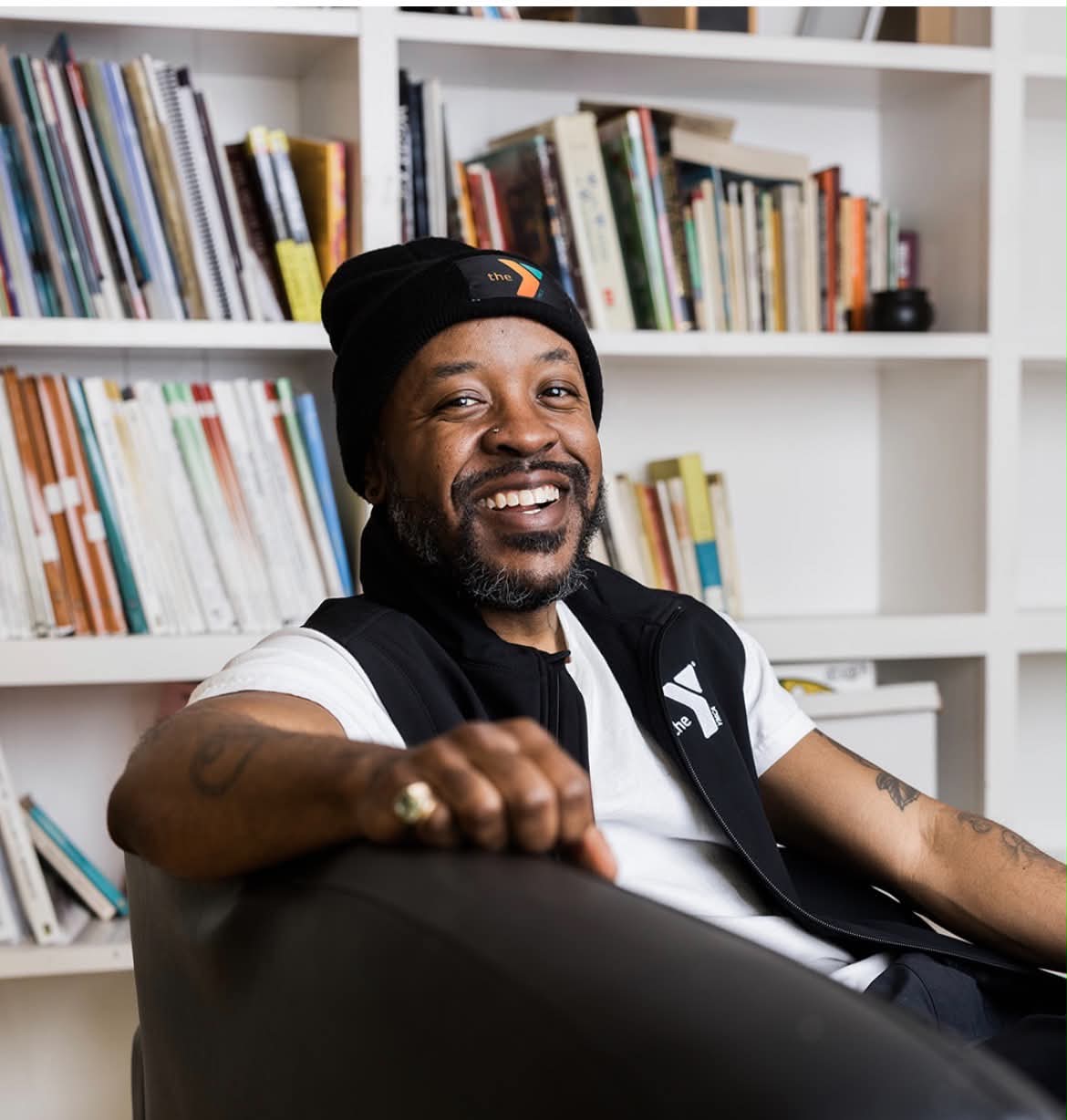 A seated person wearing a black cap and vest smiles in front of bookshelves.