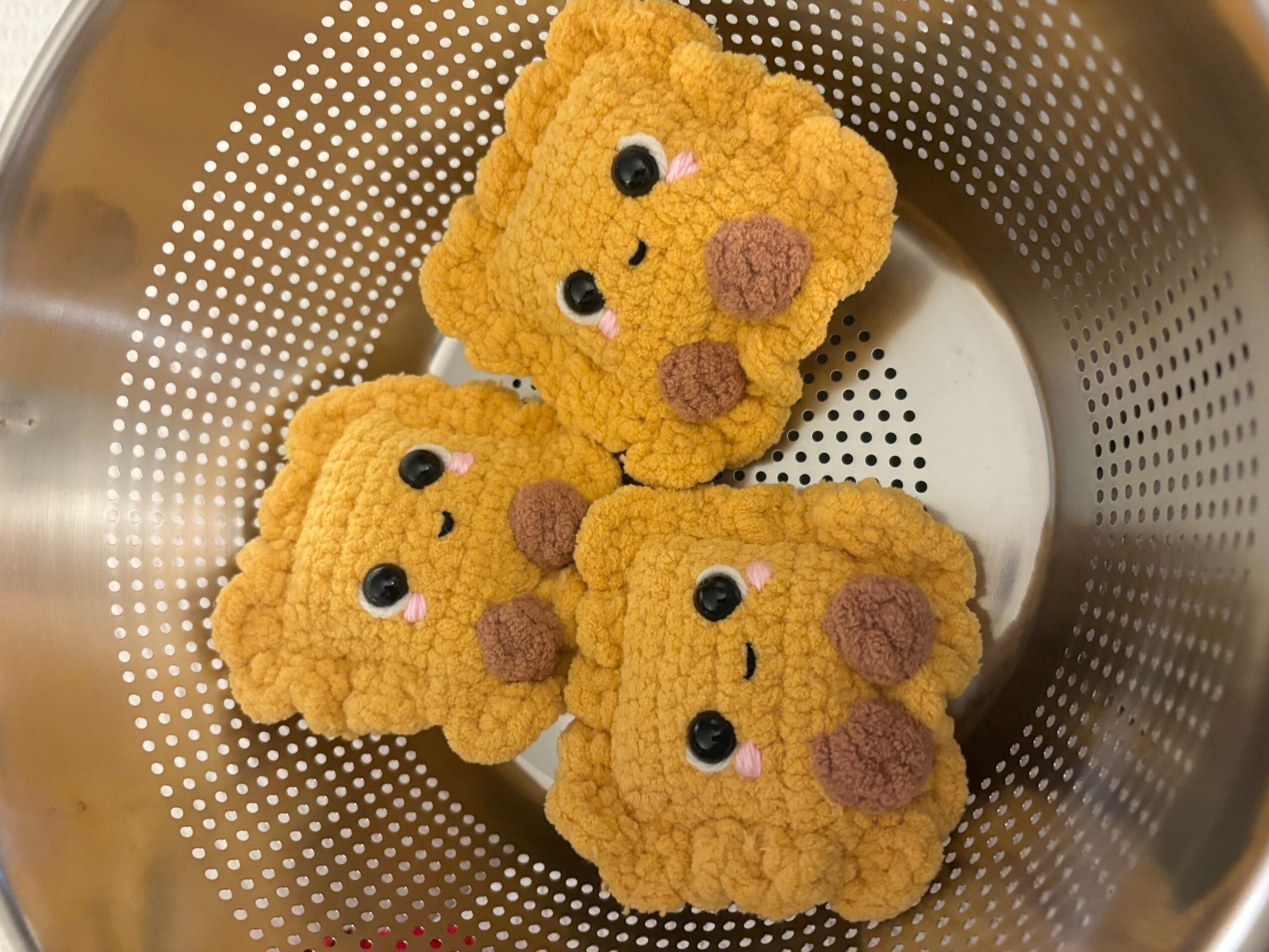 Three crocheted yellow animal-shaped items with embroidered faces sit in a perforated metal colander.