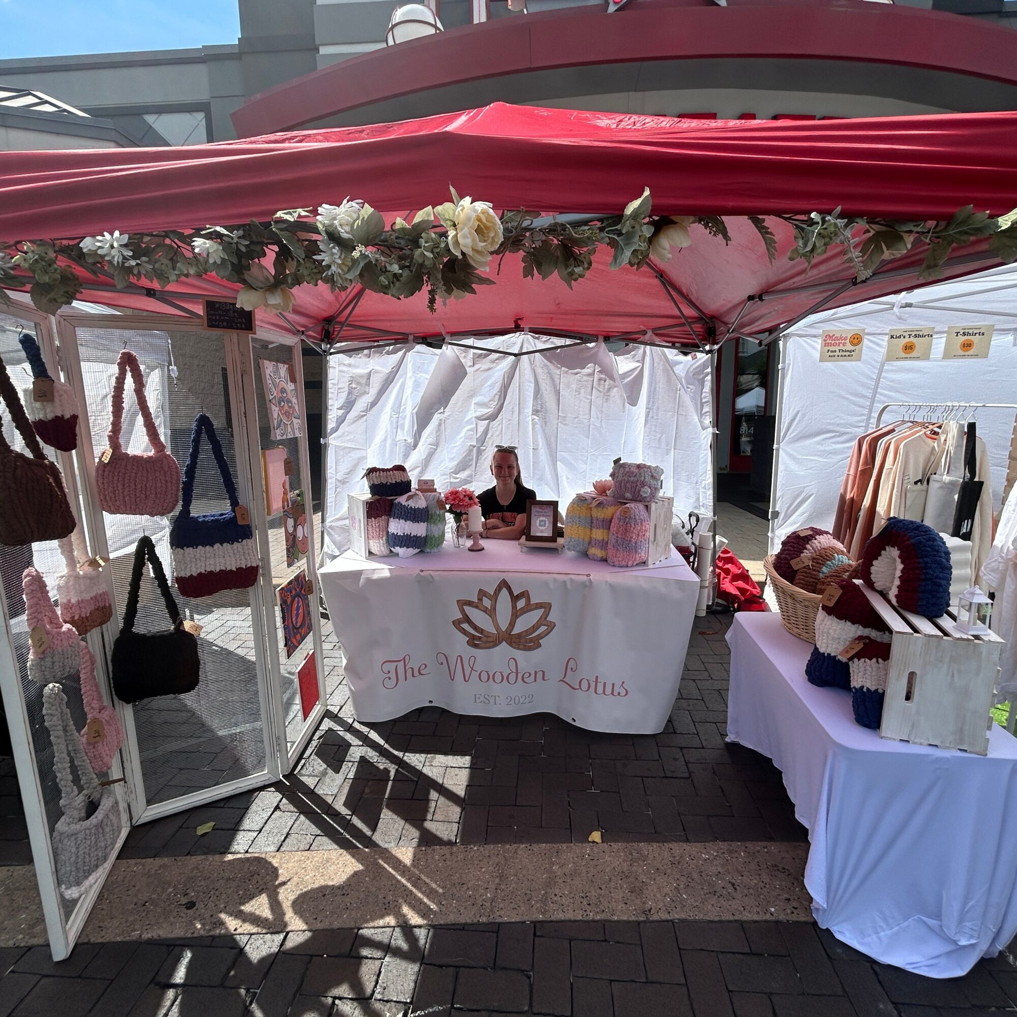 Outdoor market stall beneath a red canopy, with a white display table, flower garland, and baskets of goods.