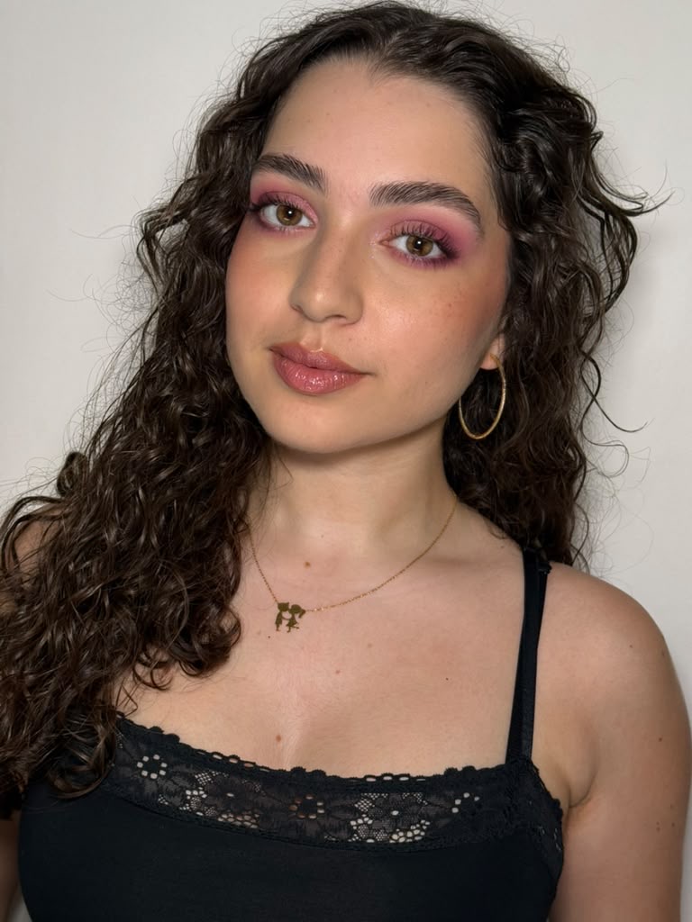 Person with curly dark hair, pink eye makeup, gold hoop earrings, and a black lace-trimmed top.