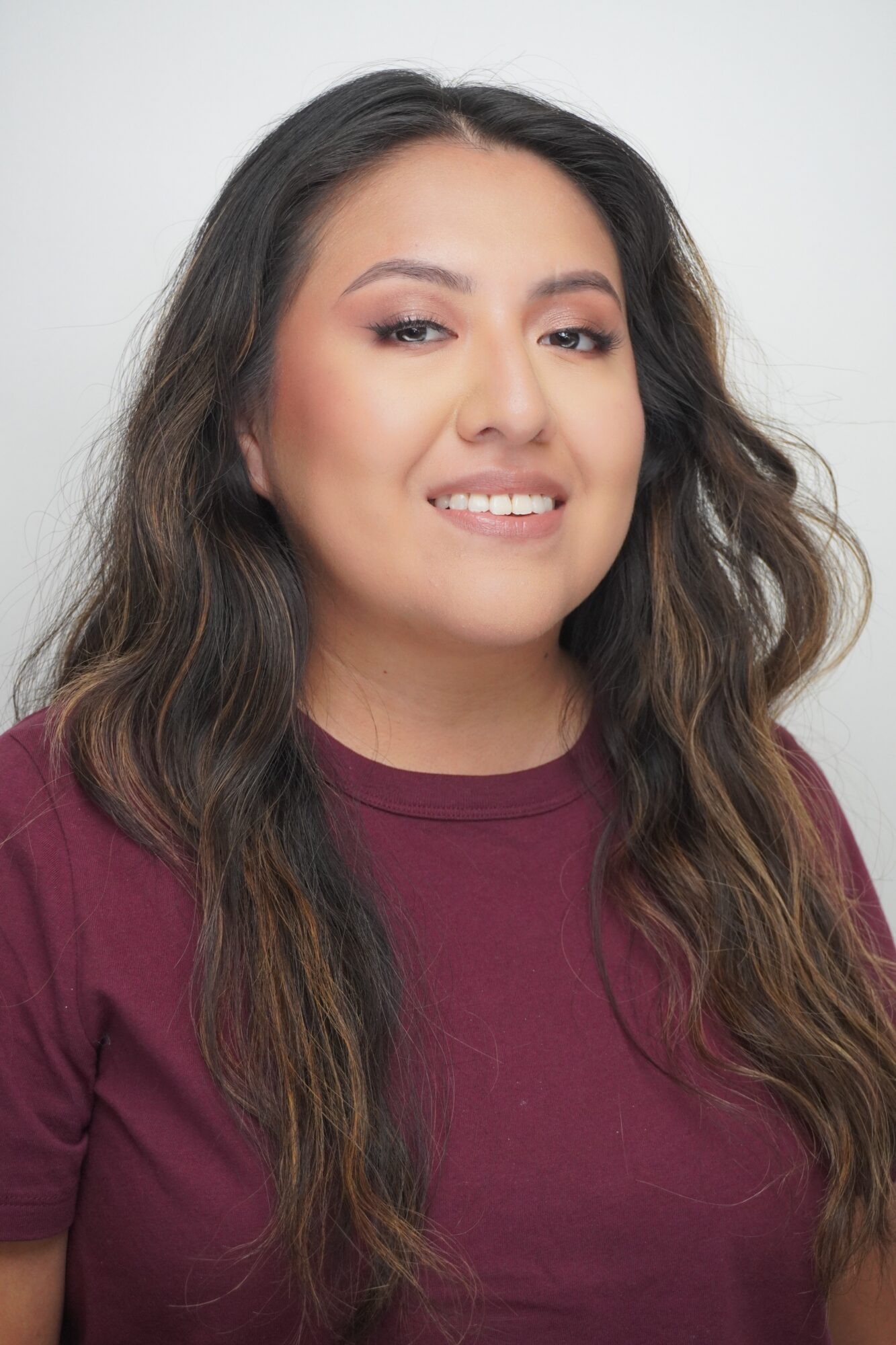 Portrait of a person with long dark wavy hair wearing a burgundy shirt against a light background.