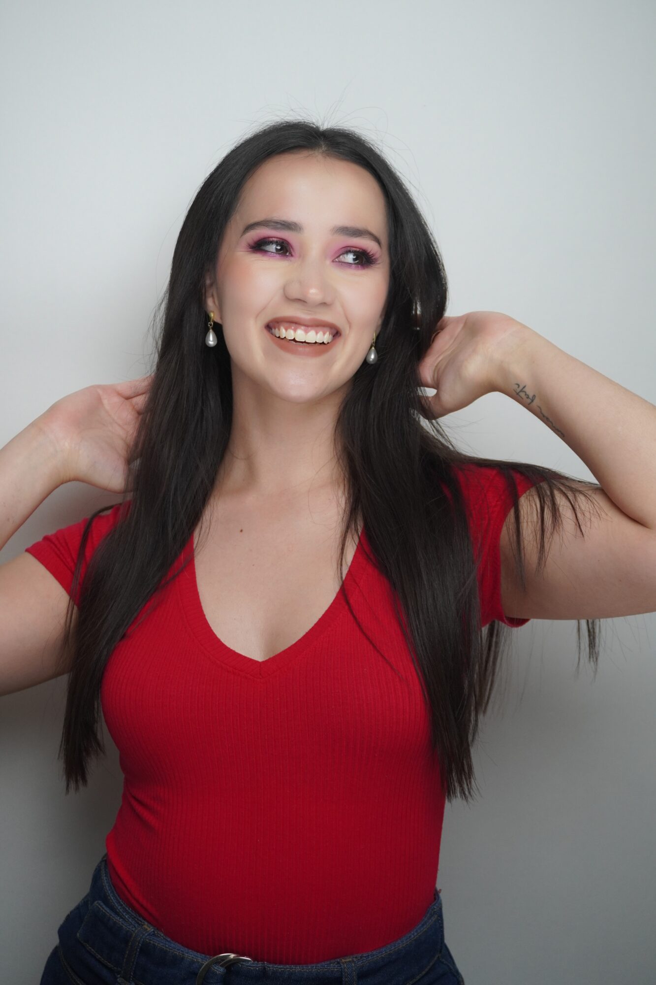 Person with long dark hair wearing a red top, smiling with both hands near her shoulders against a plain background.